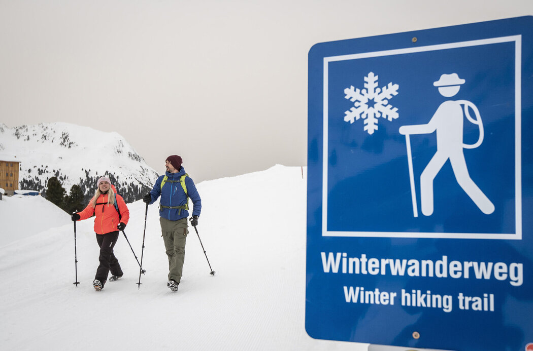 Schild von Winterwanderweg Kaiser-Franz-Josef-Weg im Kühtai im Hintergrund sind 2 Wintersportler zu sehen